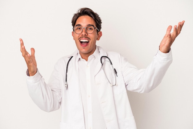 Joven médico de raza mixta aislado de fondo blanco recibiendo una agradable sorpresa, emocionado y levantando las manos.