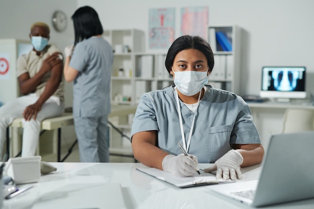 Joven médico de raza mixta en ropa de trabajo protectora rellenando documento médico por escritorio mientras su colega vacuna a un paciente masculino africano