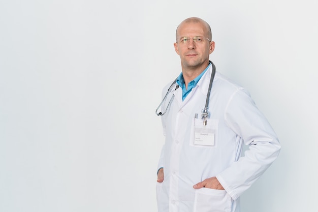 Joven médico serio en bata blanca manteniendo las manos en los bolsillos mientras está de pie junto a la pared de forma aislada