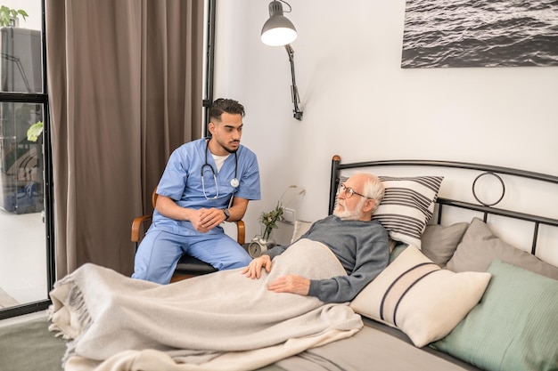 Un joven médico serio y centrado que realiza un examen visual de un anciano acostado en la cama