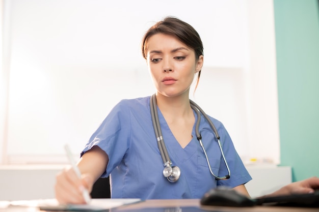 Joven médico serio o pasante en uniforme azul haciendo notas de trabajo mientras está sentado en el lugar de trabajo frente a la computadora