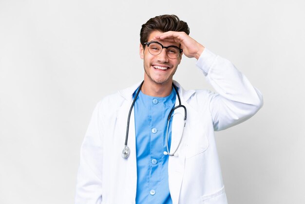 Joven médico sobre fondo blanco aislado mirando lejos con la mano para mirar algo