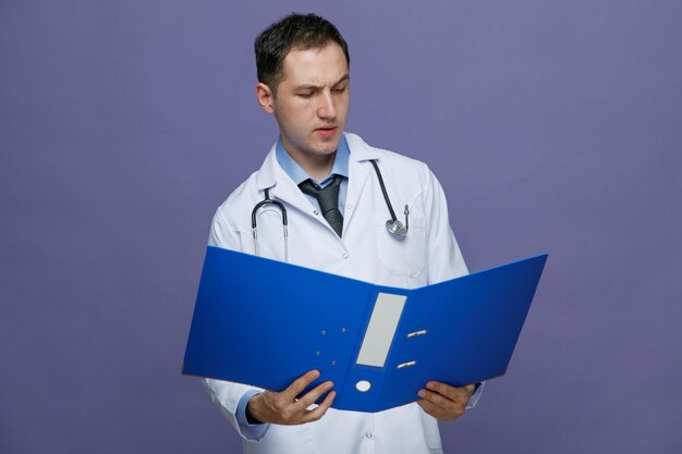 Joven médico sospechoso con bata médica y estetoscopio alrededor del cuello sosteniendo una carpeta leyendo de ella aislada en un fondo morado