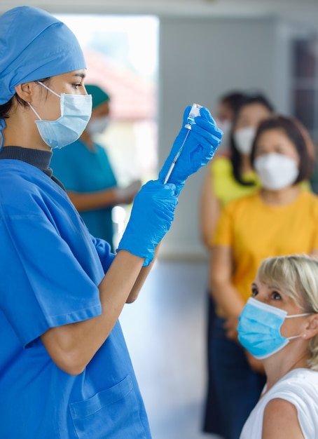 Foto el joven médico usa mascarilla, guantes de goma y uniforme de hospital azul con una jeringa de aguja que succiona la vacuna contra el coronavirus de un pequeño frasco de vidrio listo para vacunar a una paciente caucásica en la cola.