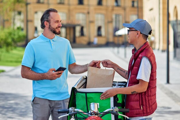 Foto joven mensajero asiático entregando comida al cliente mientras está de pie en la calle soleada