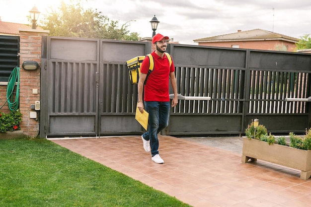 Foto joven mensajero hispano en uniforme rojo con mochila entrando a una casa privada para entregar un paquete concepto de servicio de entrega y compras en línea para pequeñas empresas