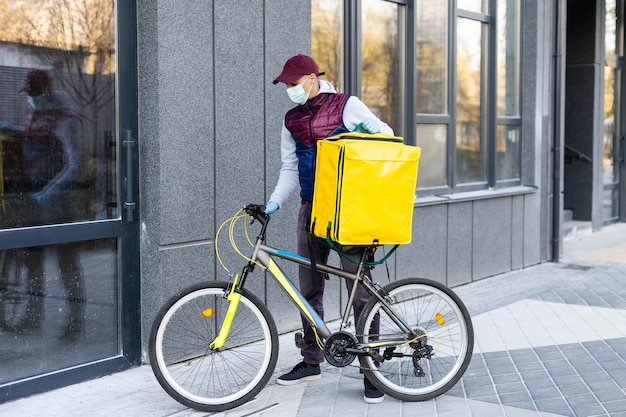 Joven mensajero con mascarilla médica entregando comida con mochila térmica amarilla, montando en bicicleta en la ciudad. Concepto de servicio de entrega de alimentos.