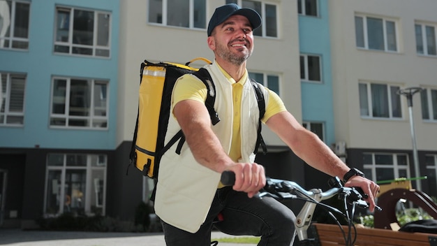 Joven mensajero con mochila térmica amarilla está haciendo una entrega del pedido a los clientes en bicicleta Concepto de entrega de alimentos