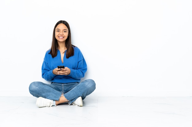 Joven mestiza mujer sentada en el piso aislado en blanco enviando un mensaje con el móvil