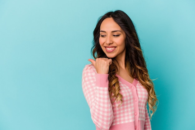 Joven mexicana aislada en la pared azul apunta con el dedo pulgar hacia afuera, riendo y sin preocupaciones.