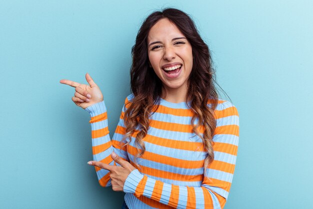 Joven mexicana aislada sobre fondo azul apuntando con el dedo índice a un espacio de copia, expresando entusiasmo y deseo.