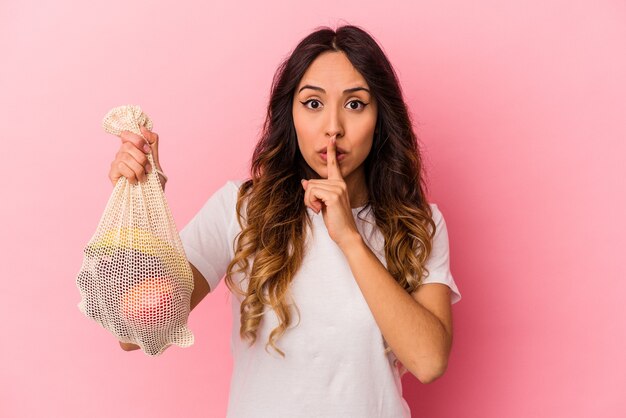 Joven mexicana sosteniendo una bolsa de frutas aislada en la pared rosa guardando un secreto o pidiendo silencio.