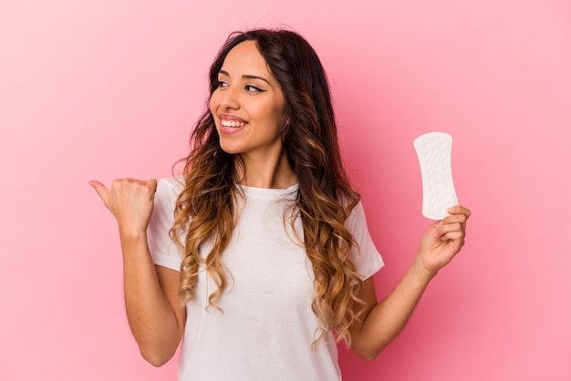 Joven mexicana sosteniendo una compresa aislada en puntos de fondo rosa con el dedo pulgar lejos, riendo y sin preocupaciones.