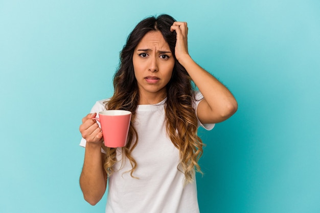 Joven mexicana sosteniendo una taza aislada en la pared azul sorprendida, ella ha recordado una reunión importante.