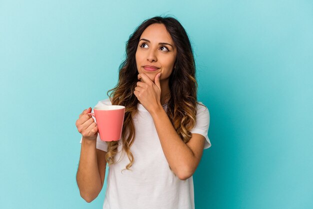 Joven mexicana sosteniendo una taza aislada sobre fondo azul mirando hacia los lados con expresión dudosa y escéptica.