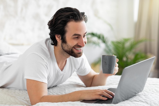 Joven milenario usando laptop en la cama bebiendo café