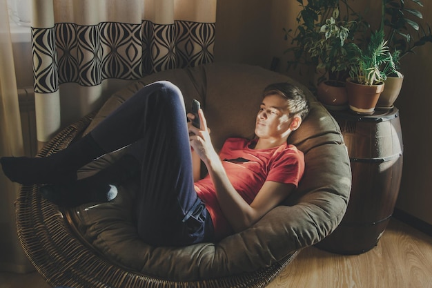 Un joven mira su teléfono inteligente acostado en una silla en casa con el resplandor del sol en su rostro