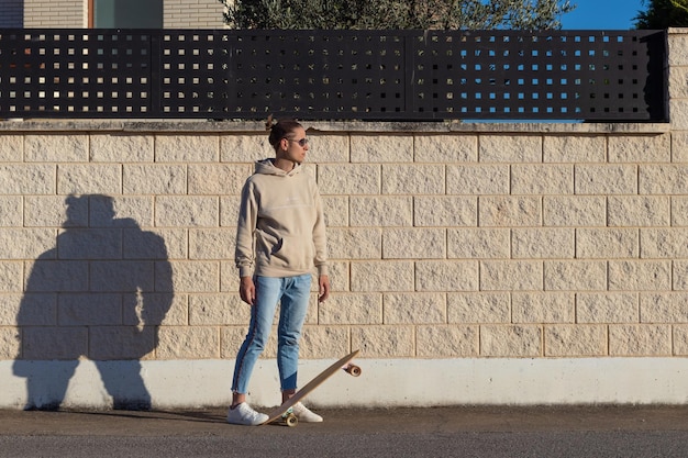 Joven mirando al sol lateral pisando una patineta contra una pared blanca a la luz de la tarde