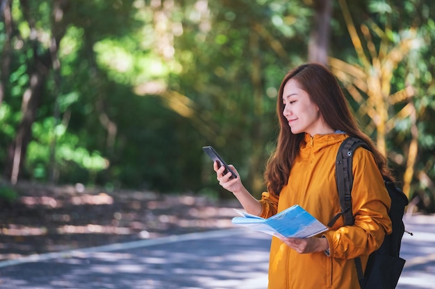 Una joven mochilera en busca de dirección en un teléfono móvil y un mapa mientras viaja por una carretera de montaña