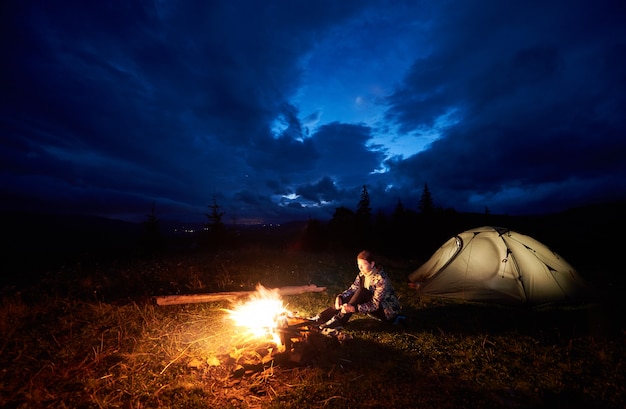 Joven mochilero disfrutando de la noche acampando en las montañas, sentado cerca de la hoguera ardiente y carpa turística iluminada bajo el hermoso cielo nublado de la tarde. Turismo, concepto de actividad al aire libre