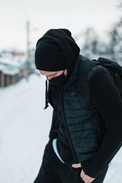 Un joven de moda con una máscara médica con ropa exterior negra con un chaleco negro, una capucha, un sombrero y una mochila camina por la calle en un día de invierno con nieve