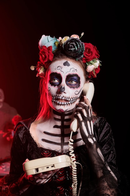 Joven modelo aterrador respondiendo una llamada de teléfono fijo, de pie sobre un fondo negro con el día de los muertos disfrazado de vacaciones. Celebrando la santa tradición mexicana con maquillaje de calavera, señora de la muerte.