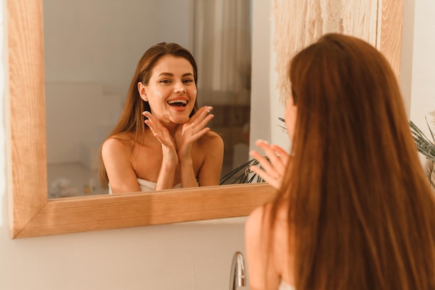 Una joven modelo riéndose aplicando crema hidratante en las mejillas en un baño moderno.