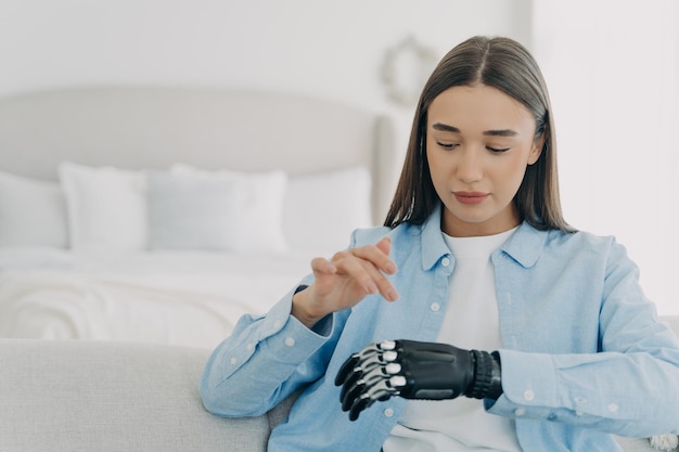 Una joven moderna con brazos artificiales entrena para usar una mano protésica biónica sentada en un sofá en casa