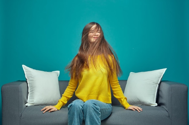Una joven moderna sacudiendo su largo cabello castaño y despeinado disfrutando del resultado del cuidado del cabello sentada en el sofá
