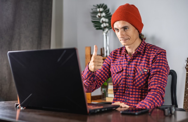 El joven moderno con una camisa a cuadros rojos y un sombrero está dando un pulgar hacia arriba mientras mira la pantalla del portátil