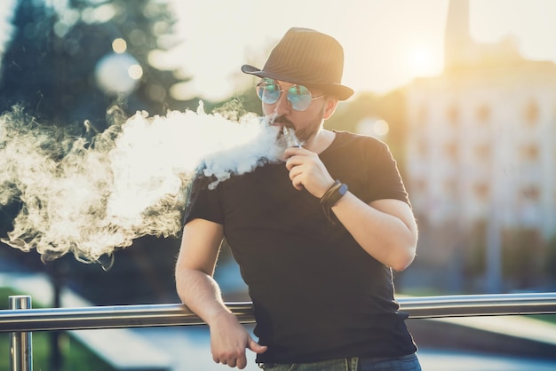 Un joven moderno y feliz con sombrero y una barba divertida Vaporizadores En el fondo, la puesta de sol sobre la imagen tonificada de la ciudad