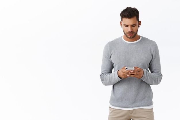 Un joven moderno con suéter gris que usa un teléfono inteligente con una expresión seria y ocupada usa auriculares inalámbricos con un mensaje de escritura en el teléfono móvil que conversa sobre negocios con compañeros de trabajo en el mensajero