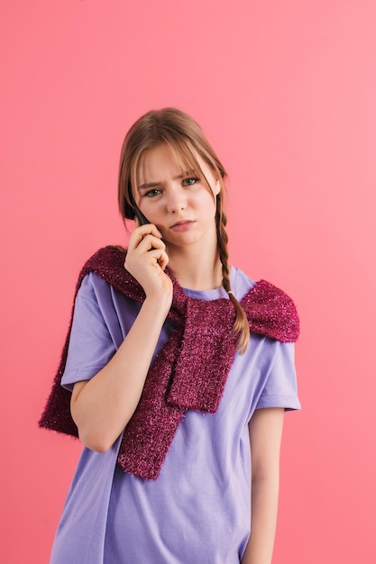 Joven molesta con dos trenzas en camiseta lila y suéter sobre los hombros hablando por teléfono celular mientras mira tristemente en cámara sobre fondo rosa