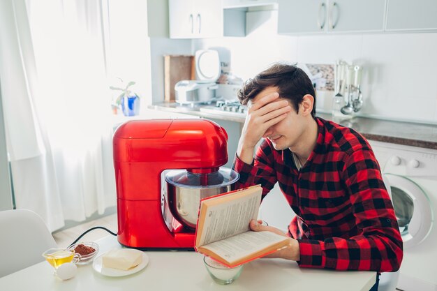 Joven molesto leyendo receta complicada en libro culinario en casa. Chico cocinando pastel con procesador de alimentos en la cocina.