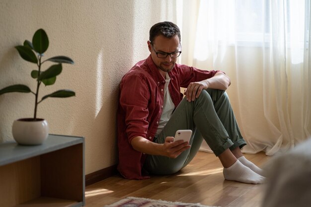 Joven molesto sosteniendo un teléfono inteligente esperando la llamada de su exnovia mientras está sentado en el suelo en casa