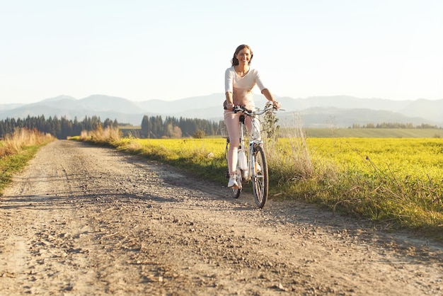 Una joven monta en bicicleta en un camino rural polvoriento, sonriendo, el sol de la tarde brilla en ella, vista desde el frente