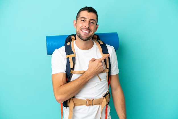 Joven montañero caucásico con una gran mochila aislada de fondo azul apuntando hacia un lado para presentar un producto