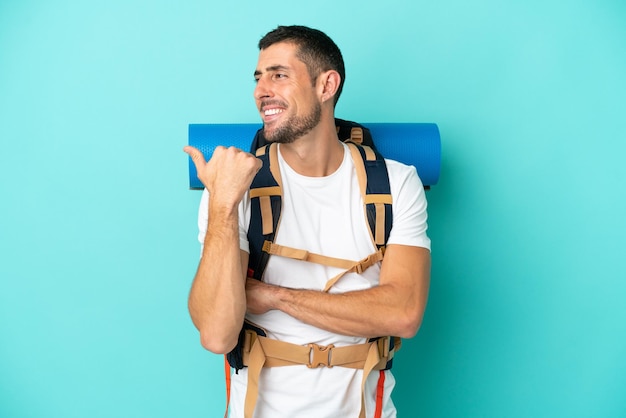 Joven montañero caucásico con una gran mochila aislada de fondo azul apuntando hacia un lado para presentar un producto