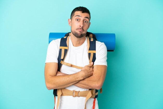 Joven montañero caucásico con una gran mochila aislada de fondo azul haciendo gestos de duda mientras levanta los hombros