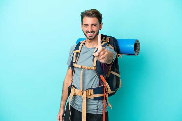 Joven montañero con una gran mochila aislada de fondo azul mostrando y levantando un dedo