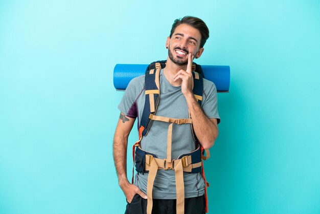 Joven montañero con una gran mochila aislada de fondo azul pensando en una idea mientras mira hacia arriba