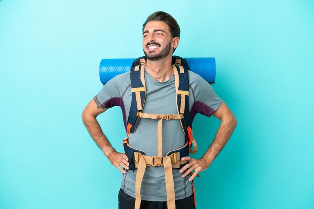 Joven montañero con una gran mochila aislada de fondo azul posando con los brazos en la cadera y sonriendo