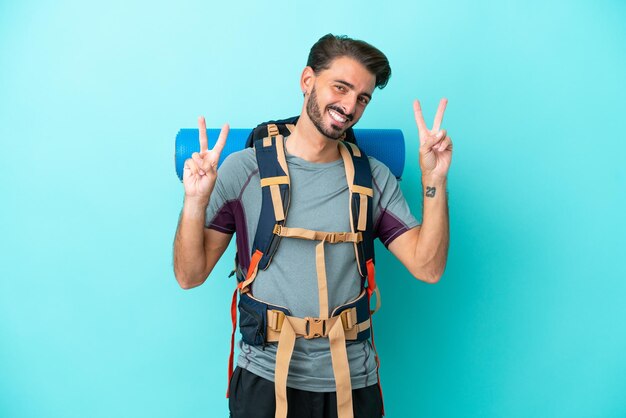 Joven montañero con una gran mochila aislada de fondo azul que muestra el signo de la victoria con ambas manos