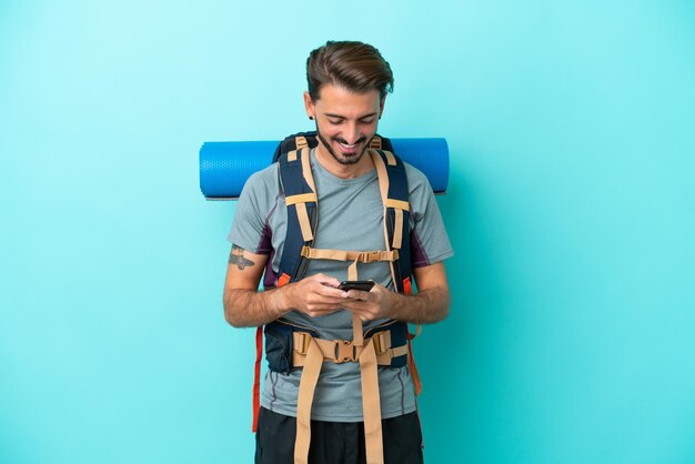 Joven montañero con una mochila grande aislada de fondo azul enviando un mensaje con el móvil