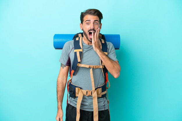 Joven montañero con una mochila grande aislada de fondo azul con expresión facial sorprendida y sorprendida