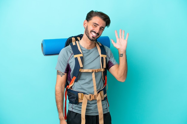 Joven montañero con una mochila grande aislada de fondo azul saludando con la mano con expresión feliz