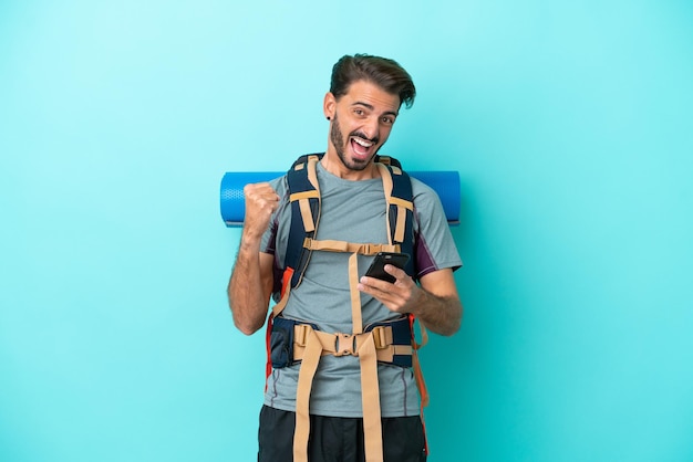 Joven montañero con una mochila grande aislada de fondo azul con teléfono en posición de victoria