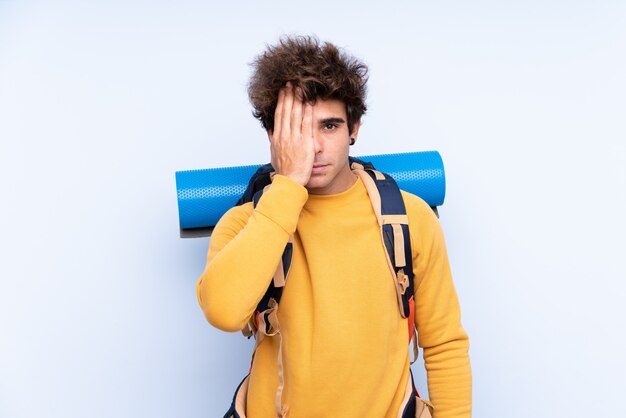 Joven montañero con una mochila grande sobre la pared azul aislada que cubre un ojo con la mano