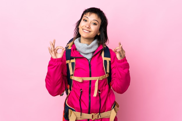 Joven montañero con una mochila grande sobre rosa aislado mostrando un signo bien con los dedos