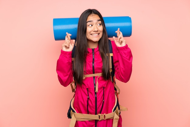 Joven montañero niña india con una mochila grande aislada en la pared rosa con los dedos cruzando